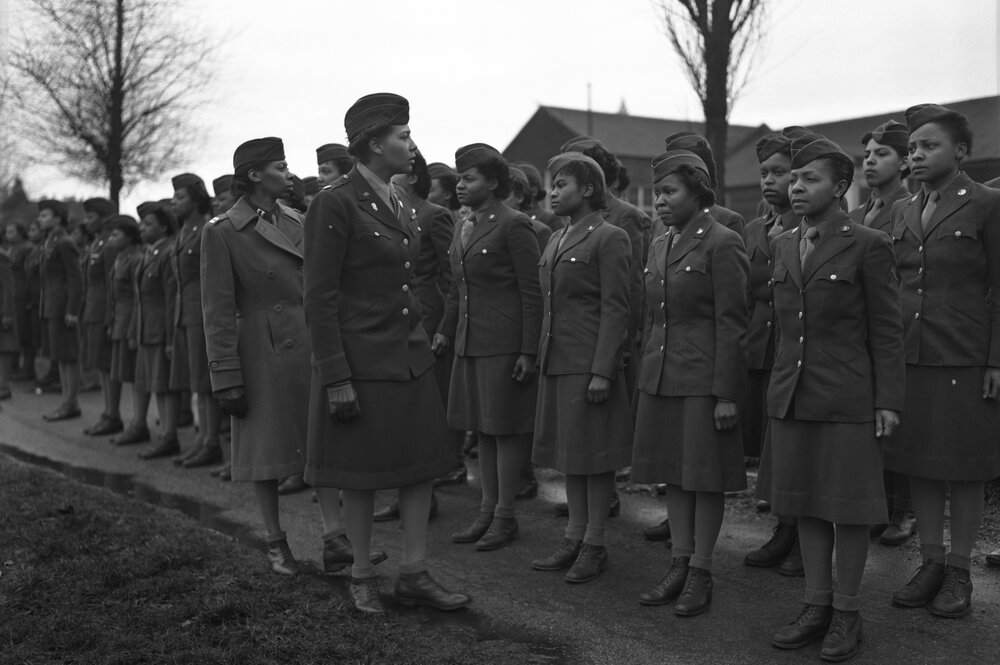 Black WACS stand at attention in two rows in uniform while their commanders walk down the line to inspect them.