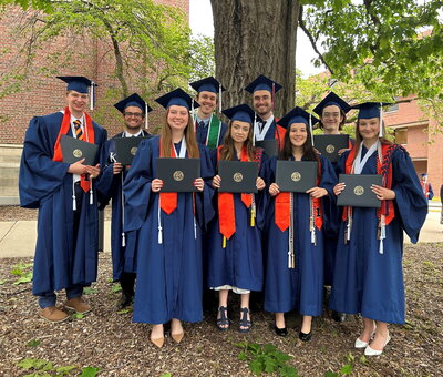 2026 Senior thesis cohort stands in a group in front of a tree in cap and gown holding their diplomas.