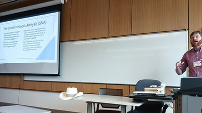 Cade Meinel stands at the front of the room near a projector screen where a slide displays info about social network analysis.