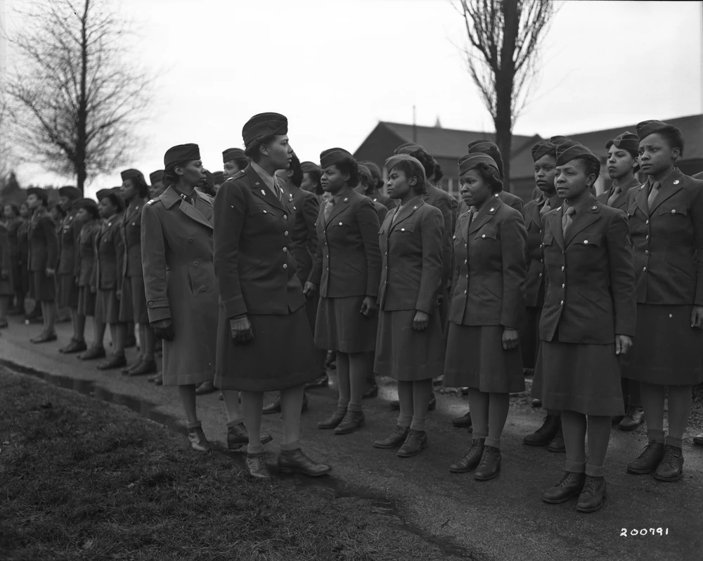 Black WACS stand at attention in two rows in uniform while their commanders walk down the line to inspect them.